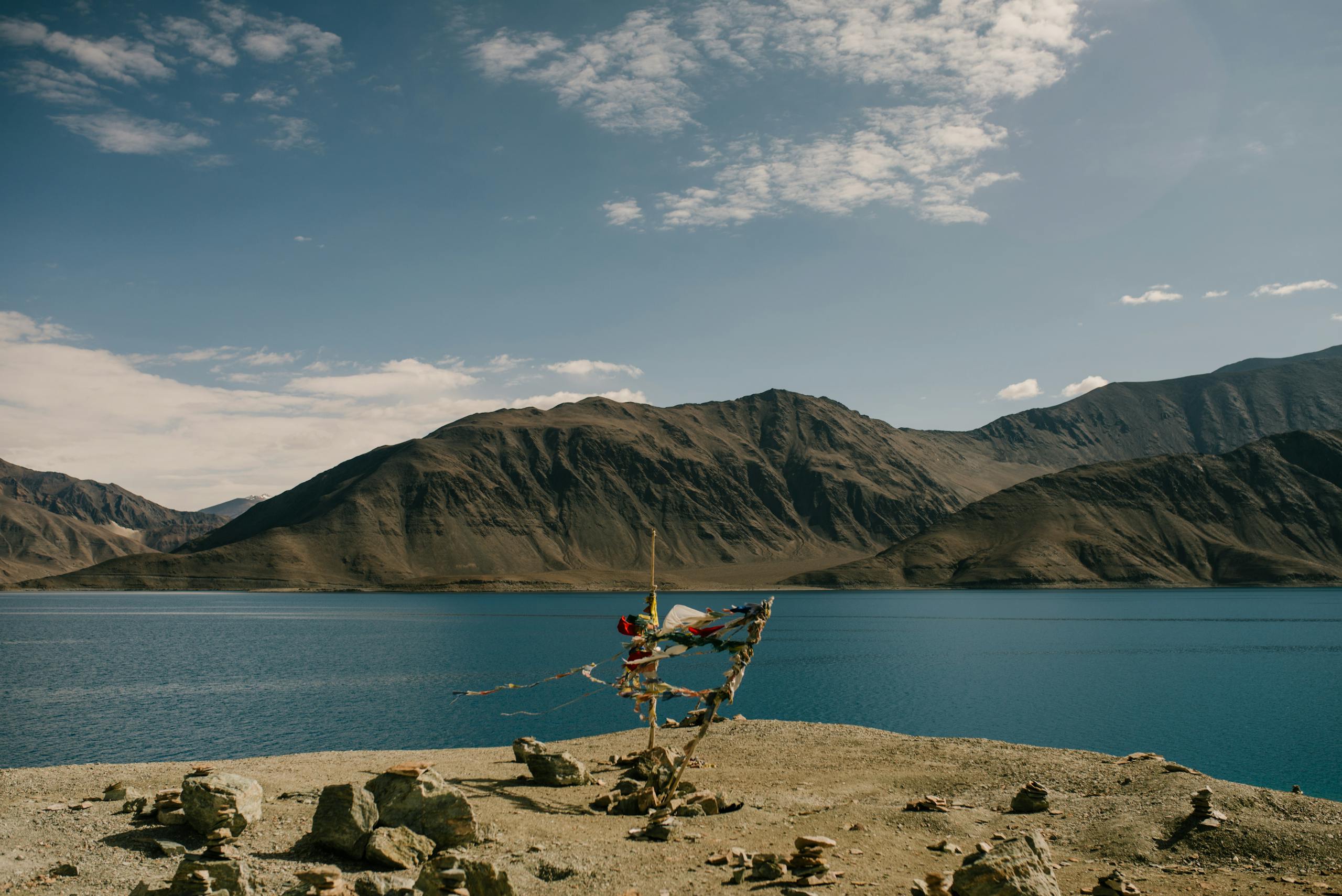 A tranquil mountain lake with Tibetan prayer flags waving in the wind, under a bright blue sky.