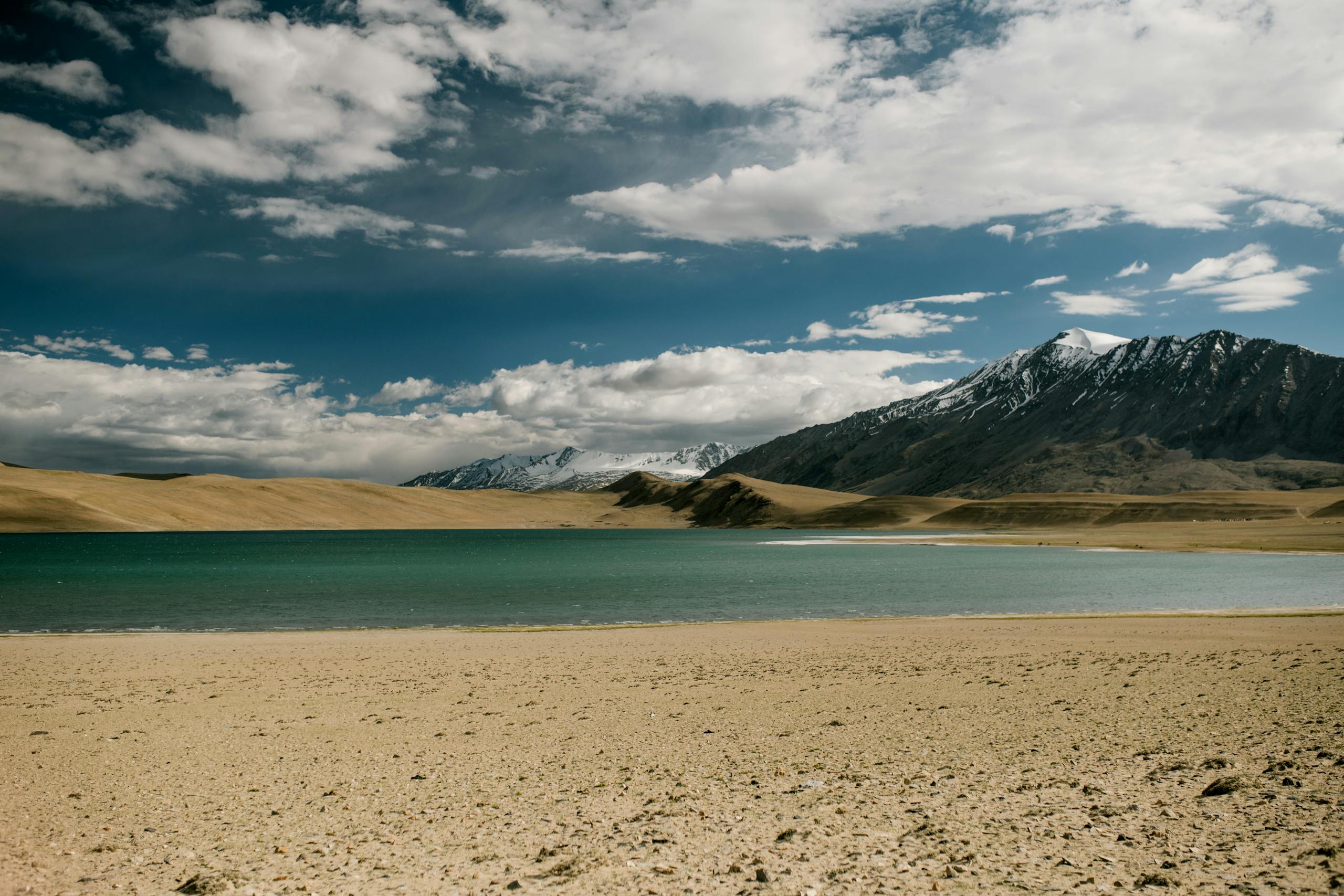 Sandy beach near river against snowy mountain peak and blue sly with clouds in nature on sunny day in countryside